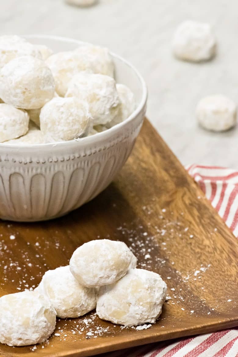 White bowl of Wedding Cookies on a wooden tray with a striped napkin on marble counter.