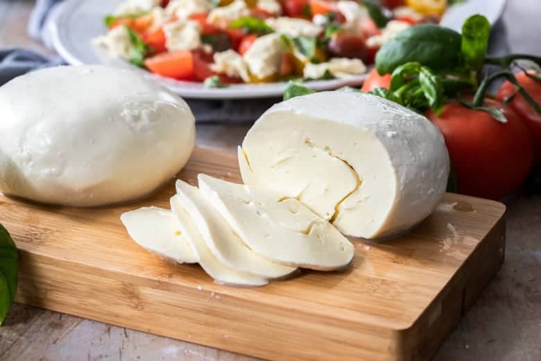 Homemade Mozzarella Cheese sliced on wooden cutting board with salad in background.