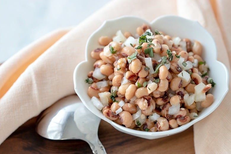 Black-Eyed Pea Salad shown in white bowl with spoon on wooden board.
