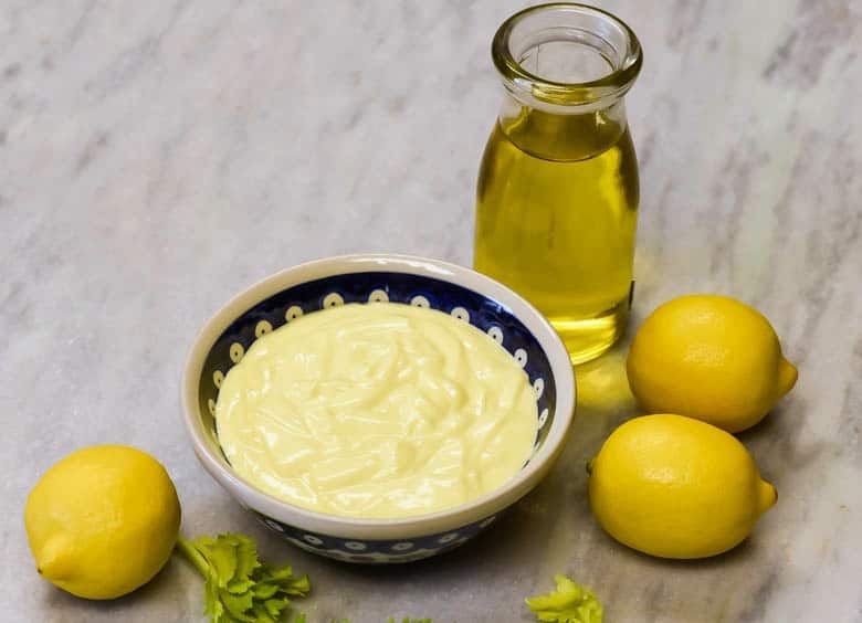 Homemade Mayonnaise recipe shown in a bowl with lemons and oil around it on a granite table.