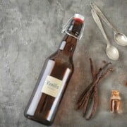 Brown bottle of homemade vanilla extract shown with vanilla beans and spoons from the post How to Make Vanilla Extract