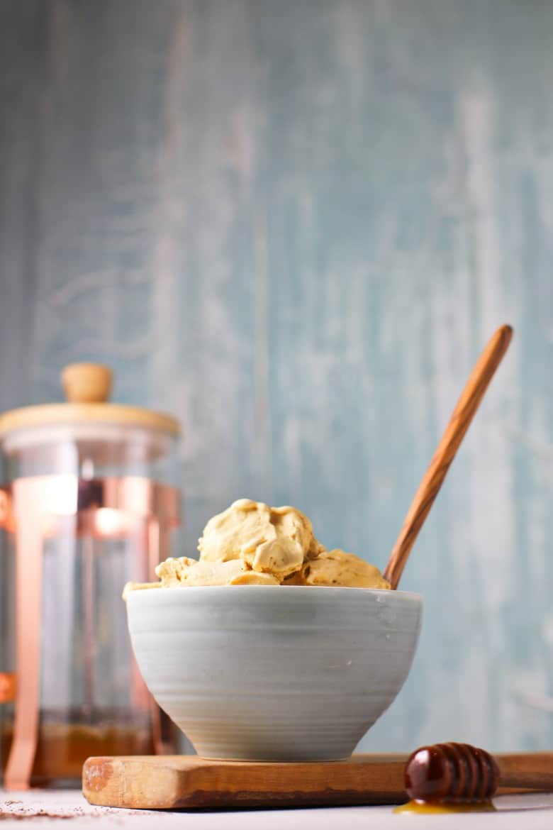 coffee ice cream in a white bowl with a wooden spoon on top of a wooden serving board with coffee in the background. 