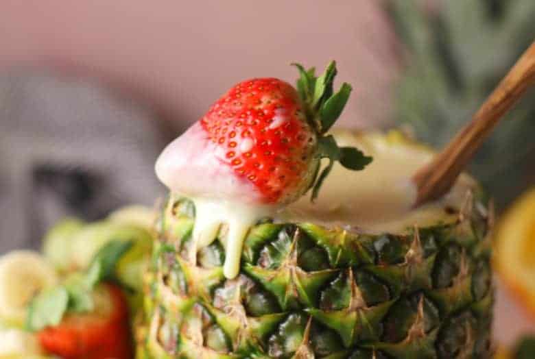 Close up of a strawberry with fruit dip recipe dripping from it on edge of a pineapple shell.