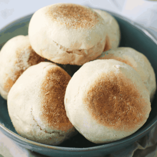 Sourdough English Muffins shown in a blue bowl on a tea towel.