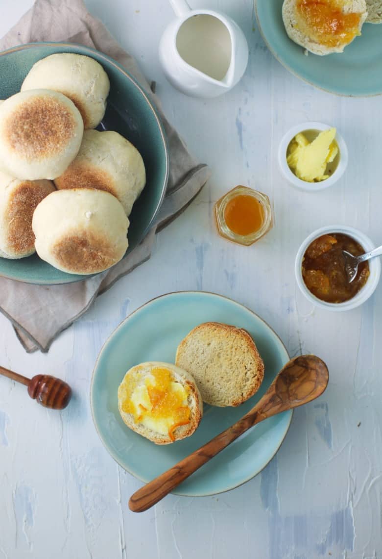 Overhead view of sourdough English muffins on a white table with jam, syrup and butter.