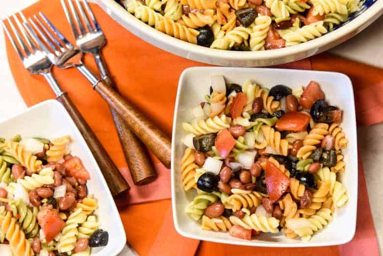 Simple Pasta Salad Recipe shown served in two white bowls and forks with serving bowl filled with salad in background