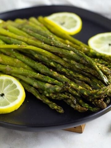Oven Roasted Asparagus shown on black plate surrounded by lemon slices.