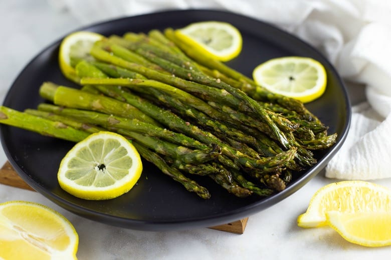 Oven Roasted Asparagus shown on black plate surrounded by lemon slices.