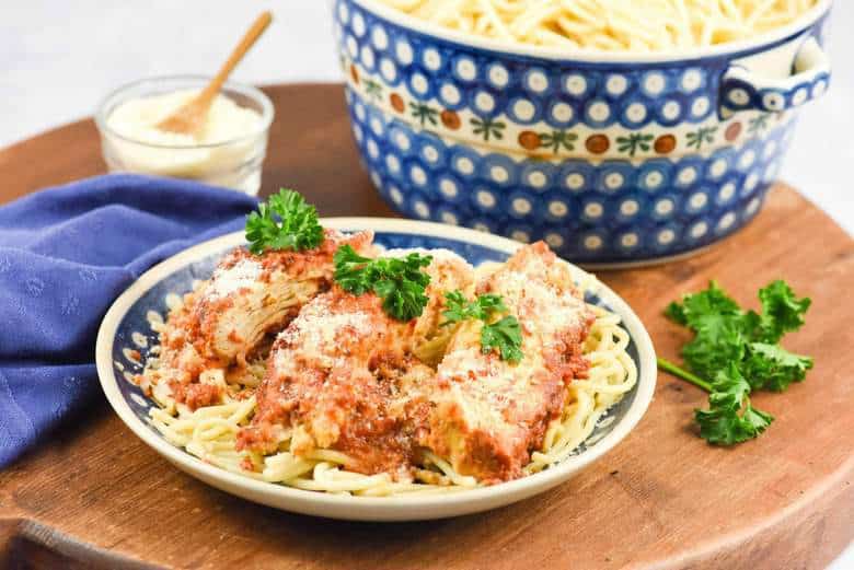 A photo of Crock Pot Chicken Parmesan on a plate on a table with a large pot of spaghetti in the background