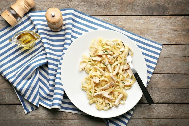 An overhead shot of Easy Chicken Alfredo on a white plate sitting on a wooden surface with a blue and white striped dishcloth