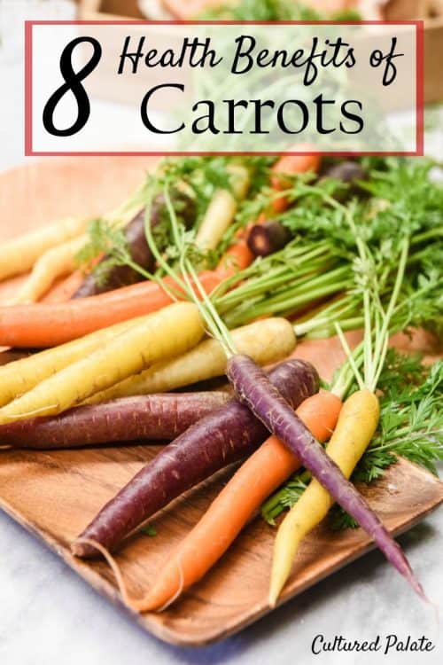 Purple, orange and yellow carrots shown on a wooden tray from the post, 8 Health Benefits of Carrots and post title at the photo top