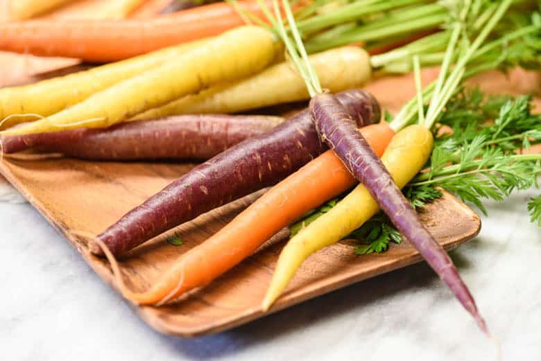 Purple, orange and yellow carrots shown on a wooden tray from the post, 8 Health Benefits of Carrots