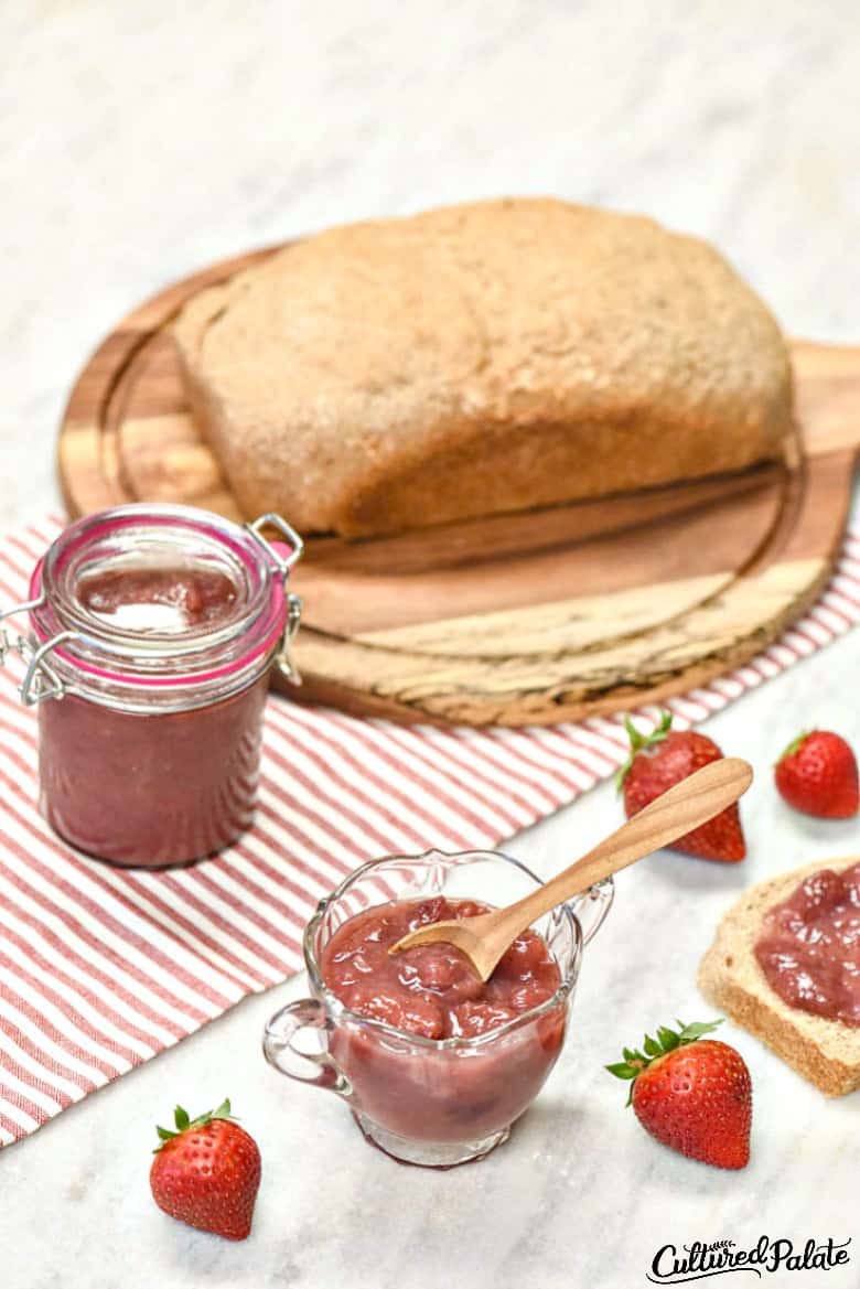 Vertical image of Instant Pot Strawberry Jam Recipe shown in a glass bowl on marble table with strawberries, a loaf of bread on a striped table runner.