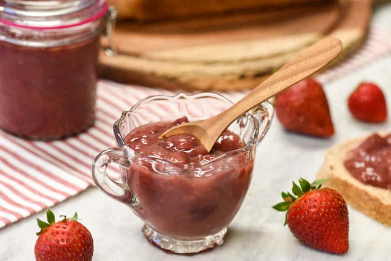 GAPS legal Instant Pot Strawberry Jam Recipe shown in a glass bowl surrounded by strawberries a glass jar with jam and a loaf of bread on wooden board in background.