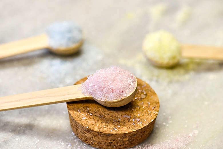 Closeup of homemade bath salts on a wooden spoon in pink, blue and yellow.