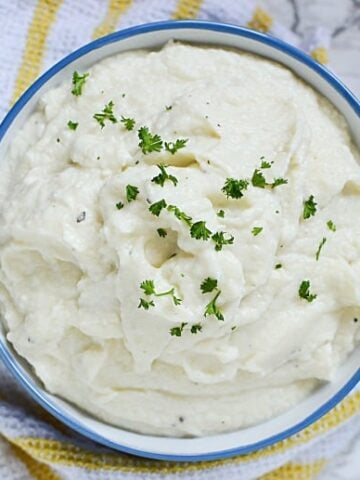 A white bowl with blue rim of mashed cauliflower from the Cauliflower Mashed Potatoes Recipe on a white and yellow striped towel