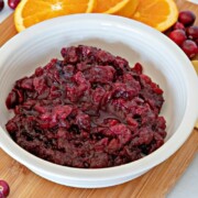 Horizontal image of a bowl of cranberry sauce on a wooden cutting board with cranberries and orange slices around it.