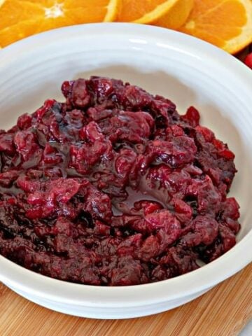 Horizontal image of a bowl of cranberry sauce on a wooden cutting board with cranberries and orange slices around it.