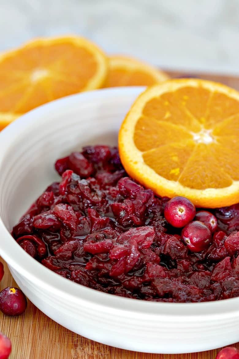 Vertical image of a bowl of cranberry sauce in a white bowl with an orange slice on top and gold spoon beside the bowl.