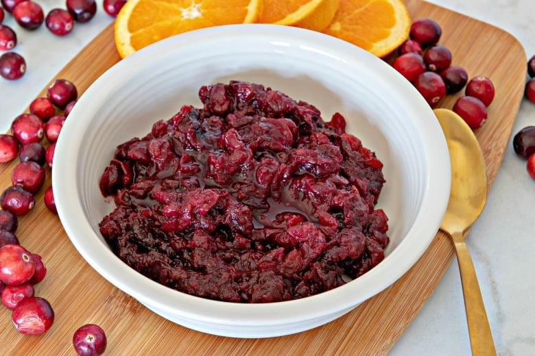 Horizontal image of a bowl of cranberry sauce on a wooden cutting board with cranberries and orange slices around it.