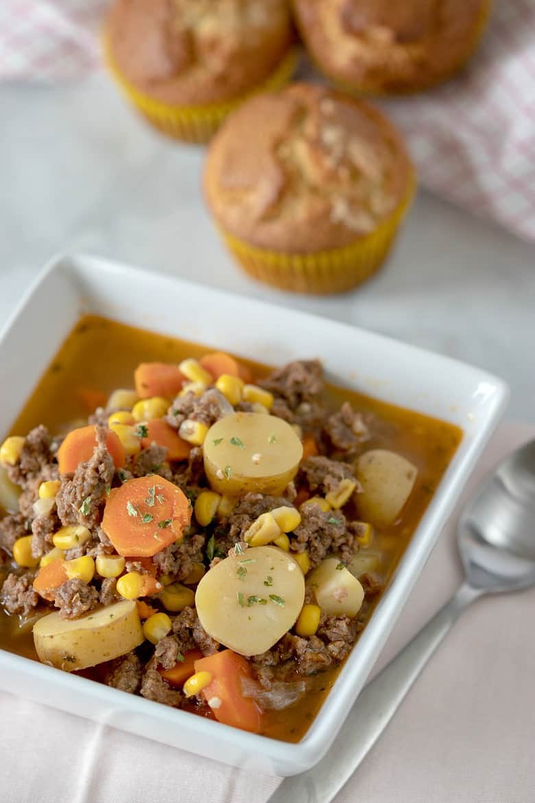 Vertical image of Instant Pot Beef Stew in a white square bowl with muffins in the background.