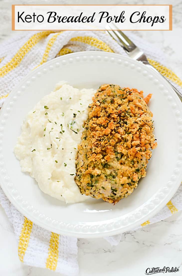 Vertical image with the title "Keto Breaded Pork Chops" showing breaded pork chop with mashed cauliflower on a white plate with yellow and white striped towel on marble background