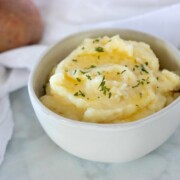 Horizontal image of Instant Pot Mashed Potatoes shown in white bowl on white marble with white napkin and potatoes on top