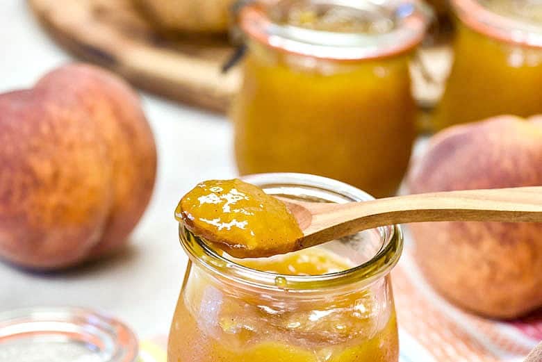 Instant Pot Peach Jam shown in a glass jar on a wooden spoon.