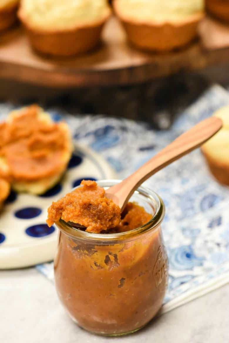 Vertical image of a spoon of pumpkin butter resting on the top of a jar with muffins in the background on blue and white napkin.