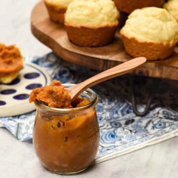Pumpkin Butter in a glass jar and spoon filled with it resting on top and muffins in the background