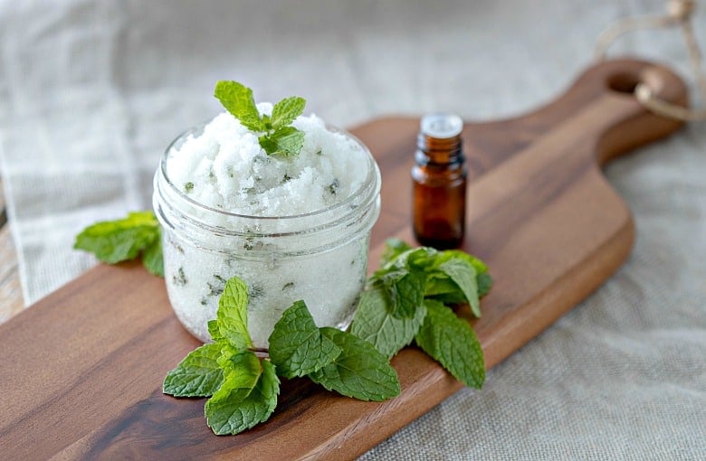 Mint Face Scrub shown in glass jar on cutting board with mint leaves around it.