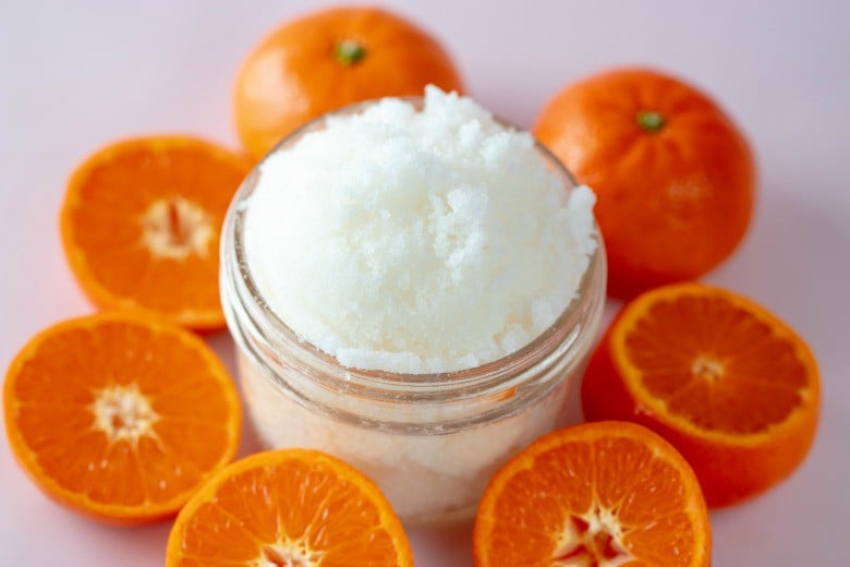 Wild Orange Sugar Scrub Recipe shown close up in glass jar with oranges around it.