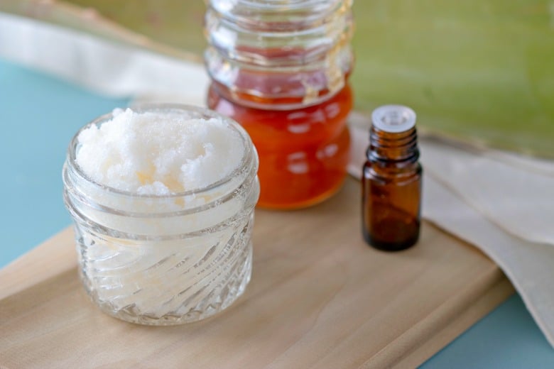 Body Scrub without Oil shown in glass jar with aloe, essential oil and honey in background on wooden board.