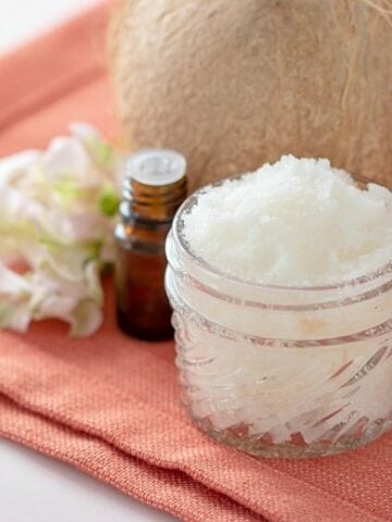 Coconut Body Scrub Recipe shown in glass jar with flowers, essential oils and coconut beside it.