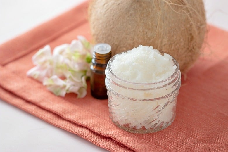 Coconut Body Scrub Recipe shown in glass jar with flowers, essential oils and coconut beside it.