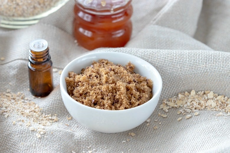 Face sugar scrub shown on linen tablecloth with oats around the bowl and honey and essential oils in background.