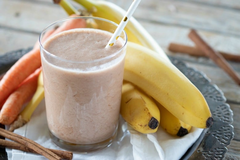 Carrot Juice with Milk shown in glass with straw and carrots and bananas on a tray.