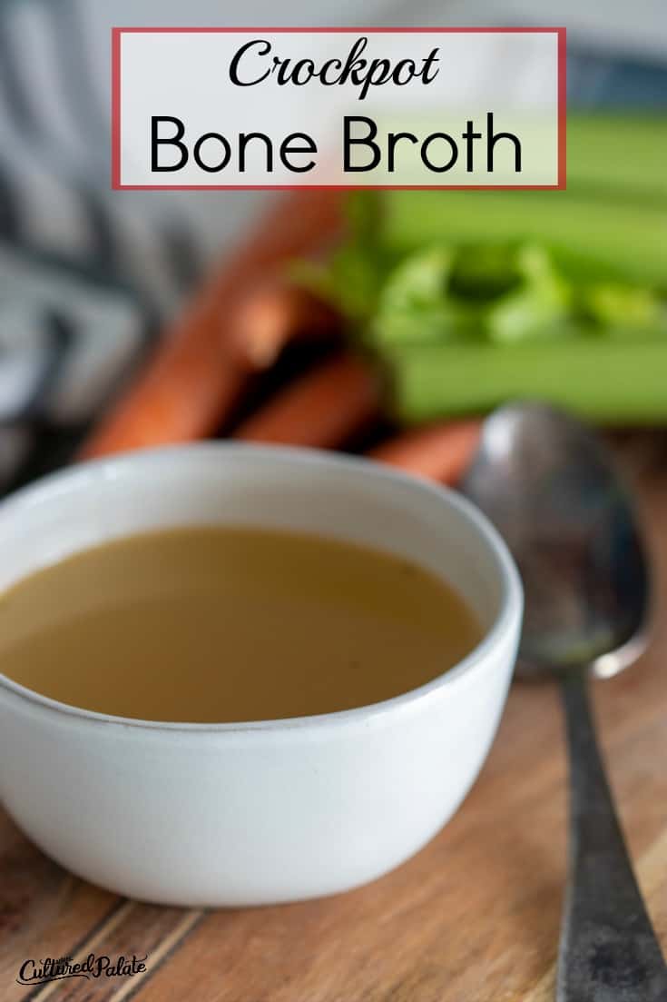 Vertical image of crockpot bone broth shown with vegetables in the background and spoon to the side of a white bowl.