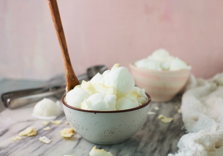 Coconut Ice Cream shown in a bowl with a wooden spoon on a marble table.